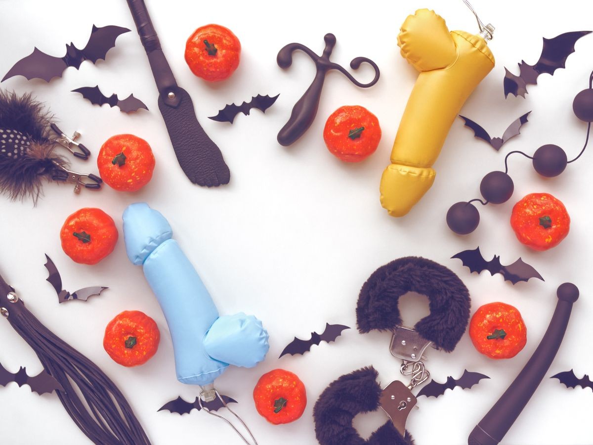 Different sex toys (spanking pad, fur handcuffs, vibrator, whip and other) are on a white background. Nearby are small pumpkins and figurines of bats. Image for advertising sex shop for Halloween.